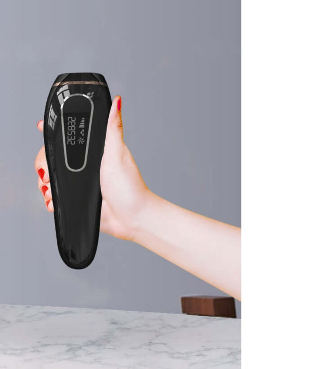 Hand holding a black permanent IPL hair removal device with LCD screen on a marble countertop.